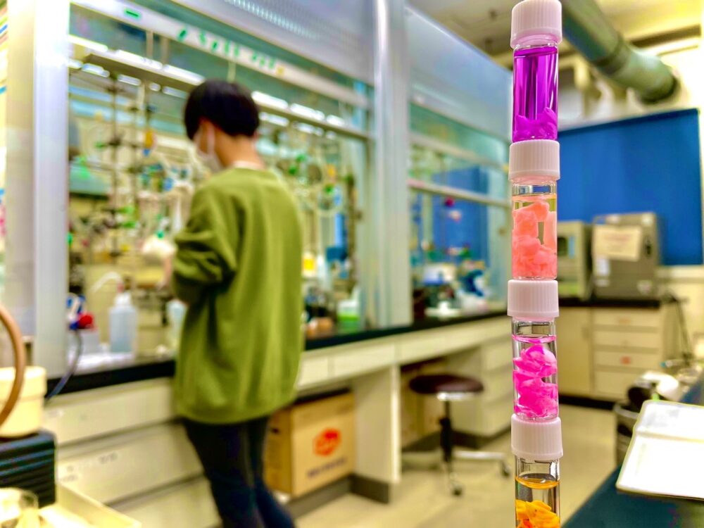 研究室の様子。学生が機器に向かって真剣に作業している。手前には、紫や桜色、ピンク色などカラフルな合成品が積まれている。