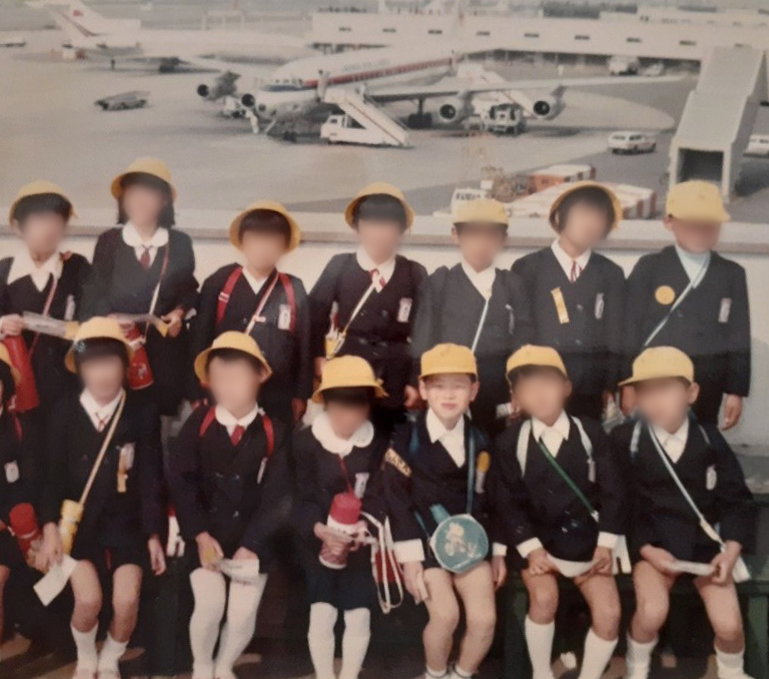 黄色のスクール帽を被り、おそろいの制服姿で集合写真を撮る、少年時代の奥村先生