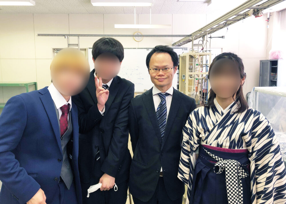 教室でスーツを着て研究室の学生と写真に写る増田先生