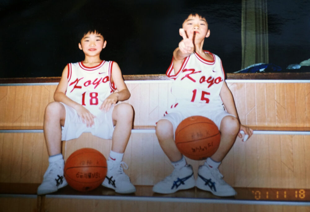 バスケットボールを足で挟み、ユニフォームを着てお兄様と並んで座る川合先生