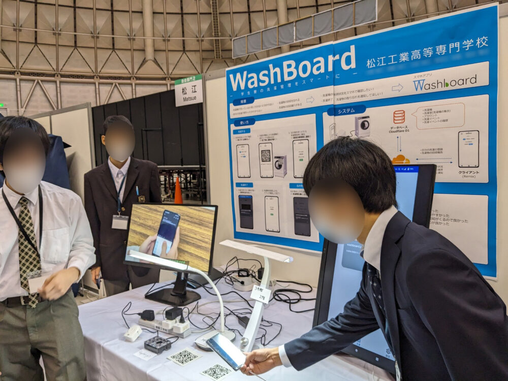 松江高専のブースにて、学生が「WashBoard－学生寮の洗濯管理をスマートに－」のアプリを説明している様子。