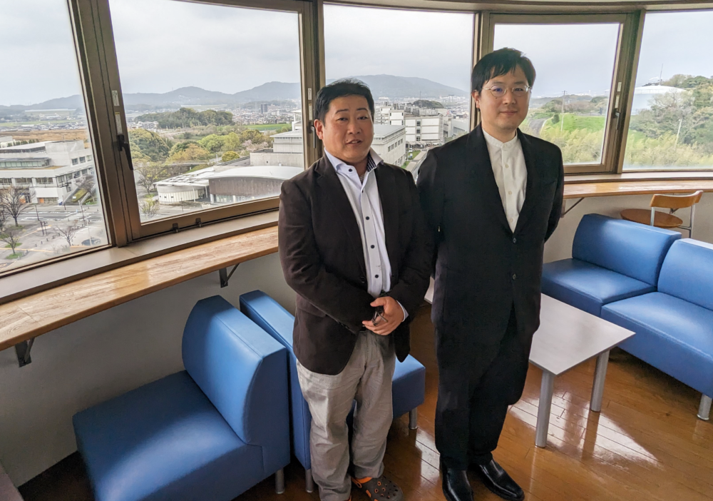 九工大の教室の窓辺で、前田先生と安川先生が並んで立ってこちらを向いている。背景の窓からは眺めの良い景色が見えている。