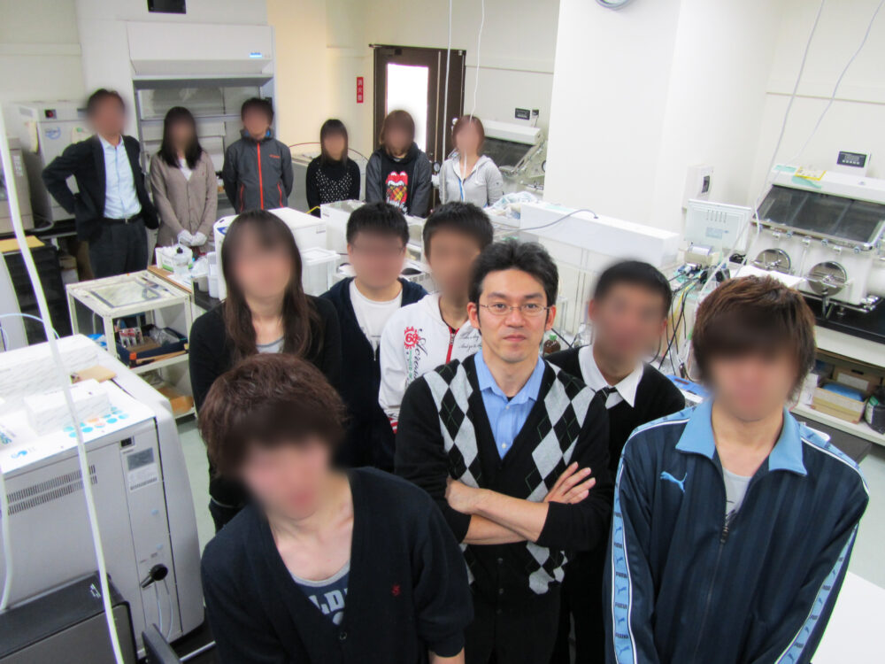 様々な機材が置かれた研究室で学生たちに囲まれた森永先生