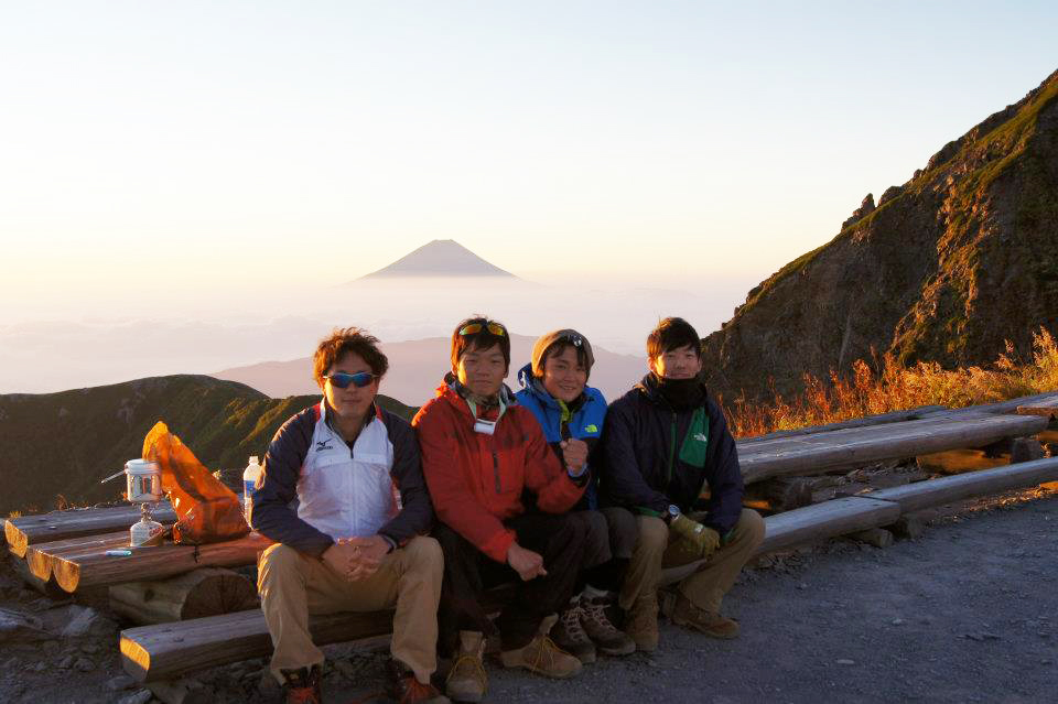 山の眺望をバックに、登山仲間とベンチに腰掛けている秋本さん