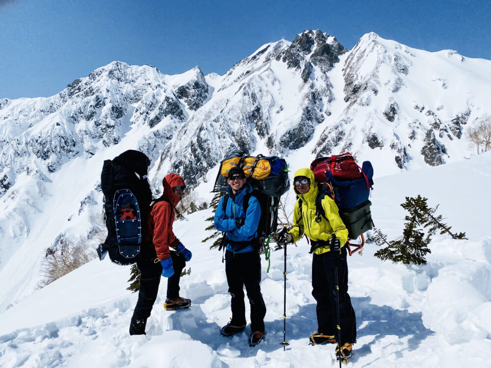 大きな荷物を背負い、
雪山の中で登山仲間と並んでいる笑顔の秋本さん