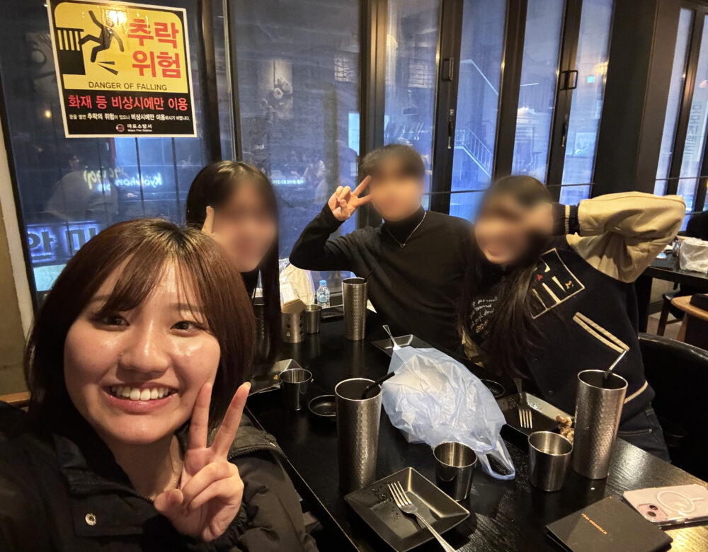 韓国の飲食店の席で、友人や案内してくれた人たちと一緒にピースサインをする巾さん