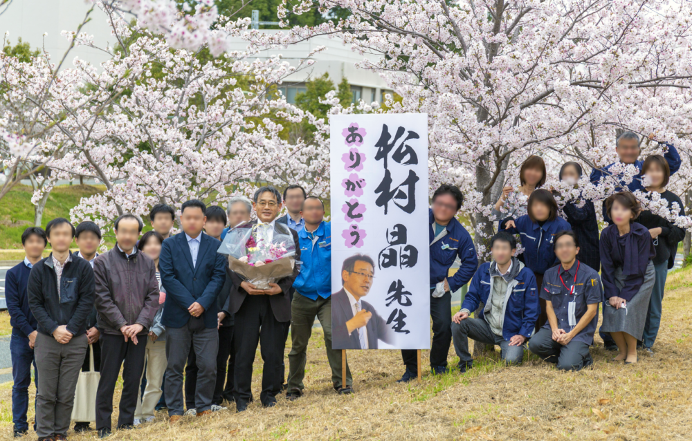 「松村晶先生ありがとう」と書かれた立て看板と一緒に、スタッフの皆さんと記念撮影