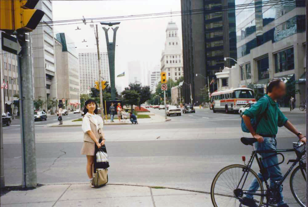 カナダの横断歩道にて一人こちらを向いている大学時代の高松先生の写真。
(備考)
大学二年生。生まれて二回目に乗った飛行機がカナダ行きの一人旅でした。
伯母の知り合いが泊めてくれるというのでカナダのトロントへ。その「伯母の知り合い」の顔も知りません。とにかく同じ方向に行きそうな人に声をかけて付いて行き、無事飛行機には乗れたものの、機上でバンクーバーでトランジットがあると判明し半泣き。見かねたカナダ人のおばさまが、自分はバンクーバーで降りるにもかかわらずトロント行きの乗り換え場所まで付いて来てくれました。しかし、おばさんはトロント行きの航空券がないので改札を通れないはず。今思えば、途中で改札の係員に詰め寄っていたから、「この子を送っていくから通して!」と交渉していたのでしょう。もしもう一度会えるなら、あのおばさんに会ってお礼を言いたいですが、どこの誰とも知れず…。その代わりに、困っている旅行者には積極的に声をかけることにしています。