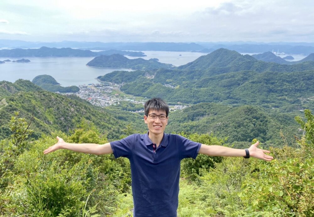 現在の下平さん。愛媛の自然を背景に手を広げて記念撮影