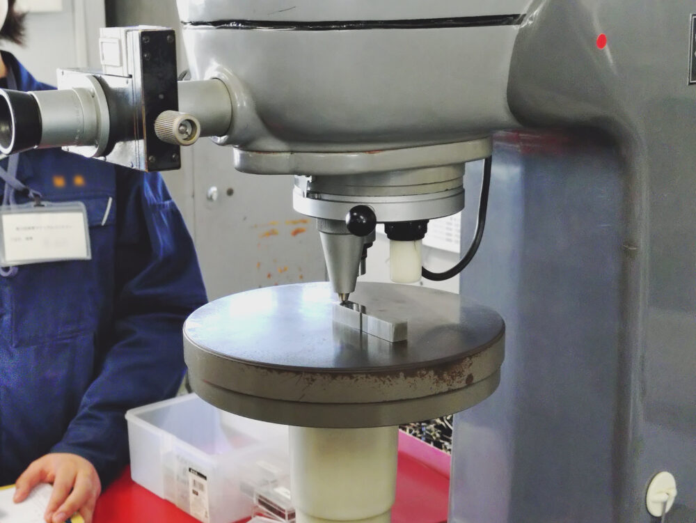 ビッカース硬さ試験の様子。正四角錐のダイヤモンド圧子を試験片に一定時間押し込む様子。